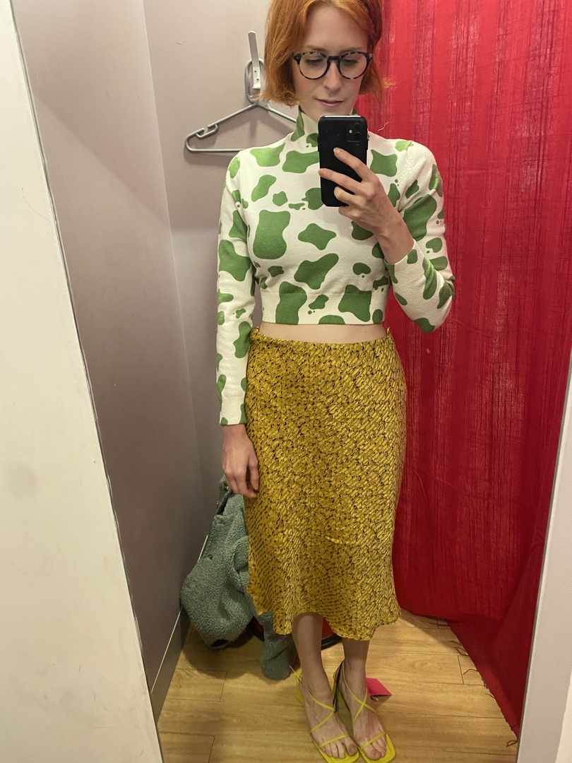 Woman in a cow-print sweater and yellow skirt trying on shoes in a changing room.