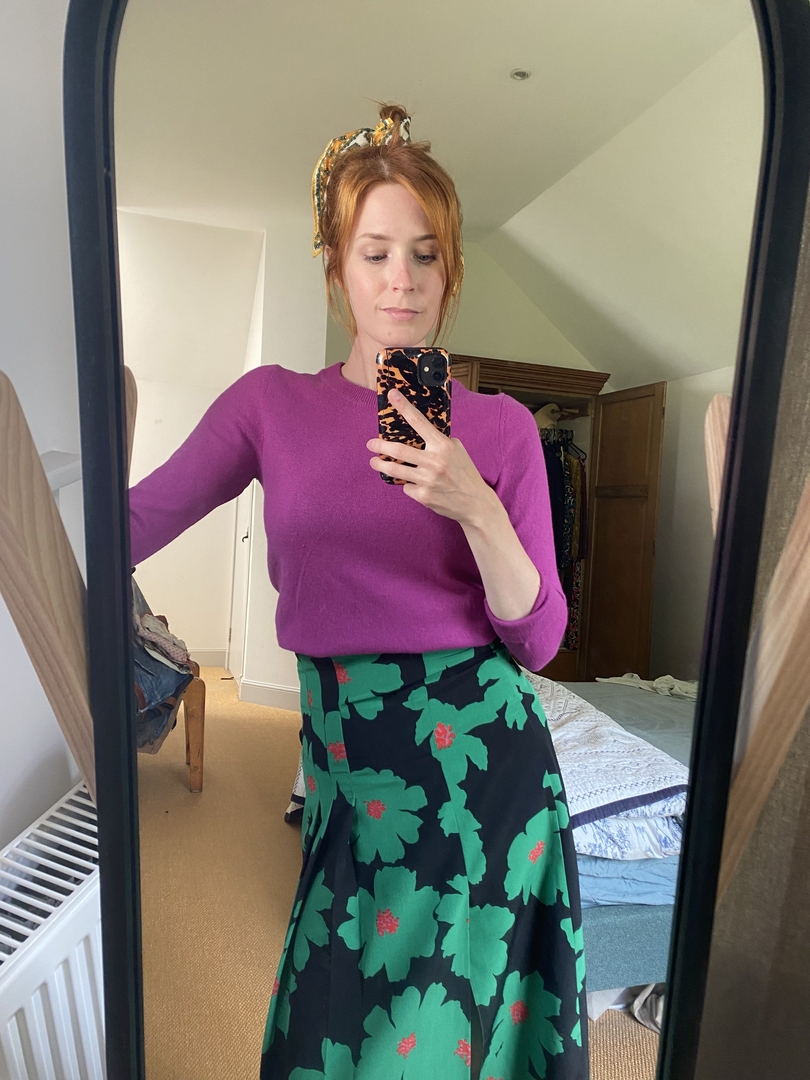 Woman in purple sweater and floral skirt taking a selfie in a mirror.